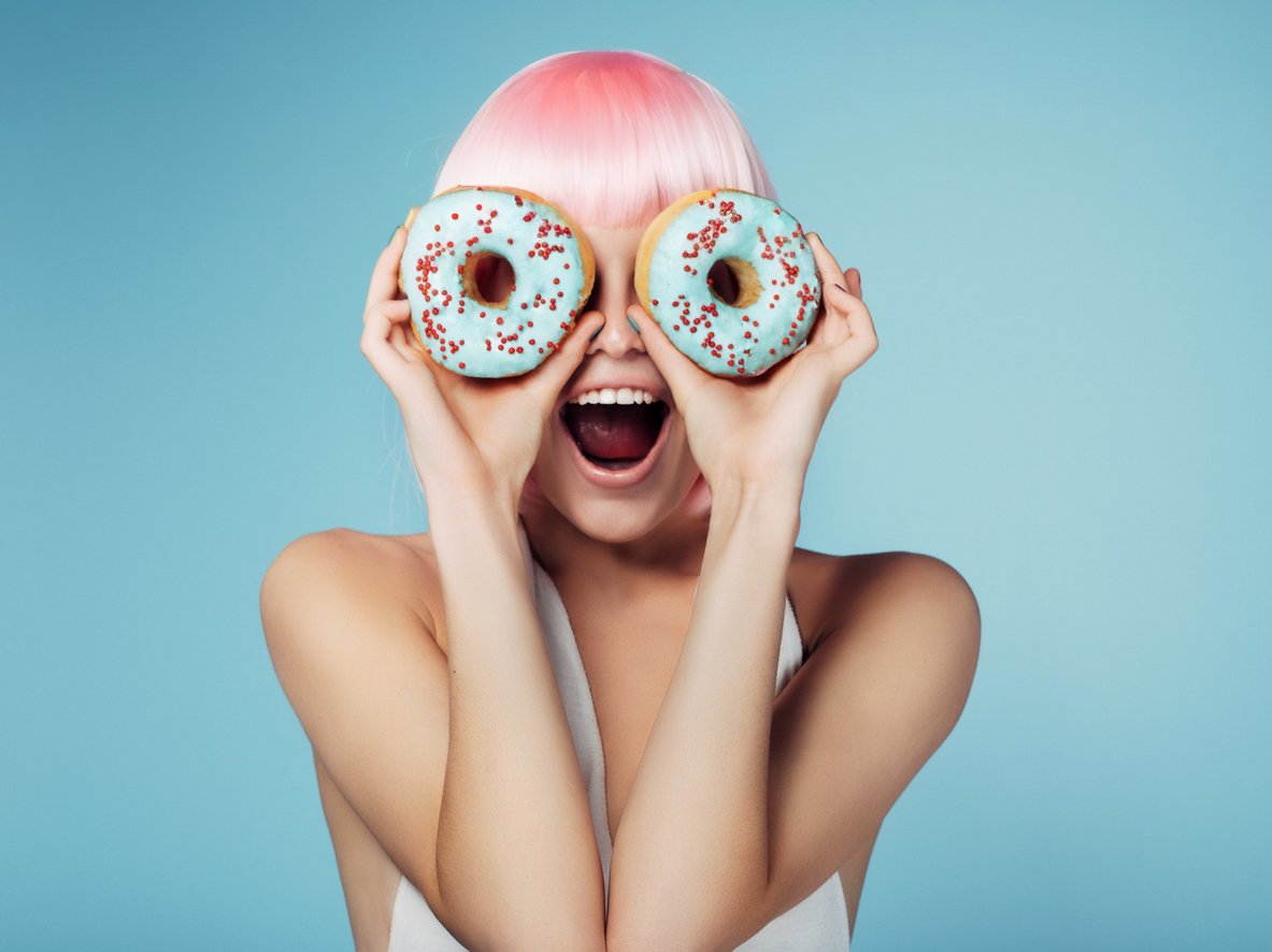 A person with a pink wig, holding up two doughnuts over their eyes, looking surprised and joyful.