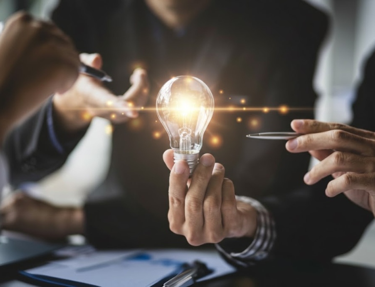 A hand offering an illuminated lightbulb during a focused business meeting, symbolizing innovative solutions.